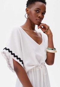 Witte jurk met brede mouwen, voorzien van zwarte scallop-rand. Touwtje in de taille, textuurstof, aangevuld met meerdere armbanden.