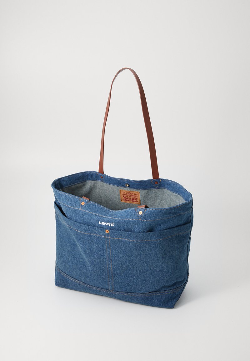 Denim boodschappentas met bruine leren hengsels, voorzien van een groot hoofdvak, een logo aan de voorkant en koperen knoopaccenten.