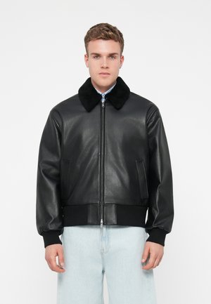 Musta nahkainen pommitakki, jossa on pehmeä musta turkiskaulus, vetoketjukiinnitys, ribattu musta hihansuu ja helma sekä kaksi sivutaskua. Suuri määrä sileää tekstiiliä.