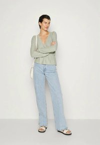 Suéter envolvente verde claro com mangas longas, jeans azul de perna larga e sandálias brancas. Modelo está de braços cruzados contra um fundo neutro.