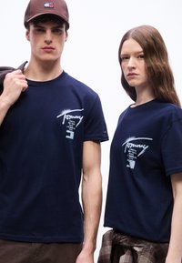 Νεαρός άνδρας και γυναίκα που φορούν μπλε σκούρα μπλουζάκια με γραφικά Tommy Jeans, με τον άνδρα να κρατάει ένα μπουφάν στον ώμο του, ουδέτερο φόντο.