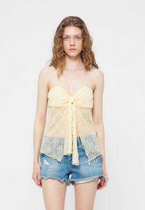Jeune femme portant un caraco en dentelle jaune transparent noué devant et un short en denim bleu déchiré, debout devant un fond blanc uni.