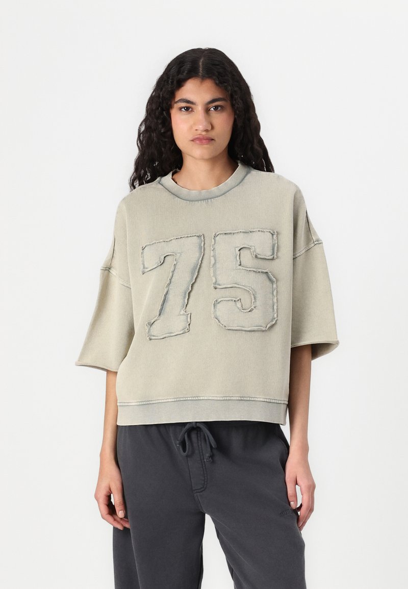 Ljusgrå oversized sweatshirt med ett stort broderat nummer "75" på framsidan. Mjuk textur, korta ärmar och rund halsringning.