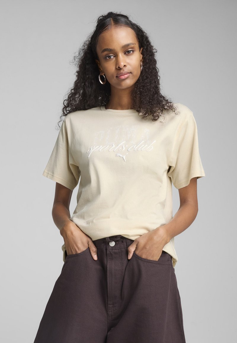 Beige Baumwoll-T-Shirt mit kurzen Ärmeln, das eine weiße Grafik mit der Aufschrift "PUMA Sports Club" und einem kleinen Logo zeigt. Getragen mit dunkelbraunen Hosen.