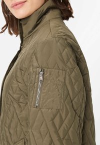 Quilted olivgrüne Jacke mit hohem Kragen, ausgestattet mit einer Reißverschluss-Tasche auf dem oberen Ärmel und einer weichen, glatten Textur.