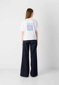 T-shirt blanc à manches courtes avec un graphisme bleu et rose "Calvin KLEIN" au dos. Assorti à un jean en denim large et foncé.