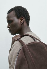 Zaino in suede marrone con texture morbida e forma arrotondata. Accentuato da spallacci in pelle liscia. Il modello indossa un maglione marrone chiaro.