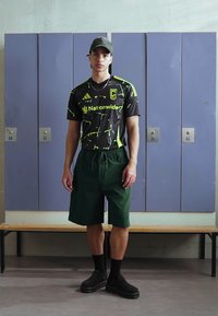 Camiseta deportiva negra y verde neón con patrón abstracto, pantalones cortos verdes, calcetines negros y zapatos negros sin cordones. El fondo presenta taquillas.