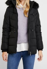 Chaqueta acolchada negra con capucha de piel sintética. Cuenta con bolsillos con cremallera, puños acanalados y un diseño acolchado. Llevada sobre un suéter de punto lila claro.