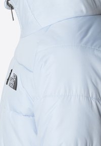 Jasnoniebieska kurtka ocieplana z kapturem i czarnym logo The North Face na klatce piersiowej, z widocznymi detalami pikowania.