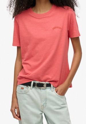 Osoba ubrana w koralowy t-shirt z krótkim rękawem z małym logo na piersi oraz jasnoniebieskie dżinsy z czarnym paskiem i ozdobną klamrą.