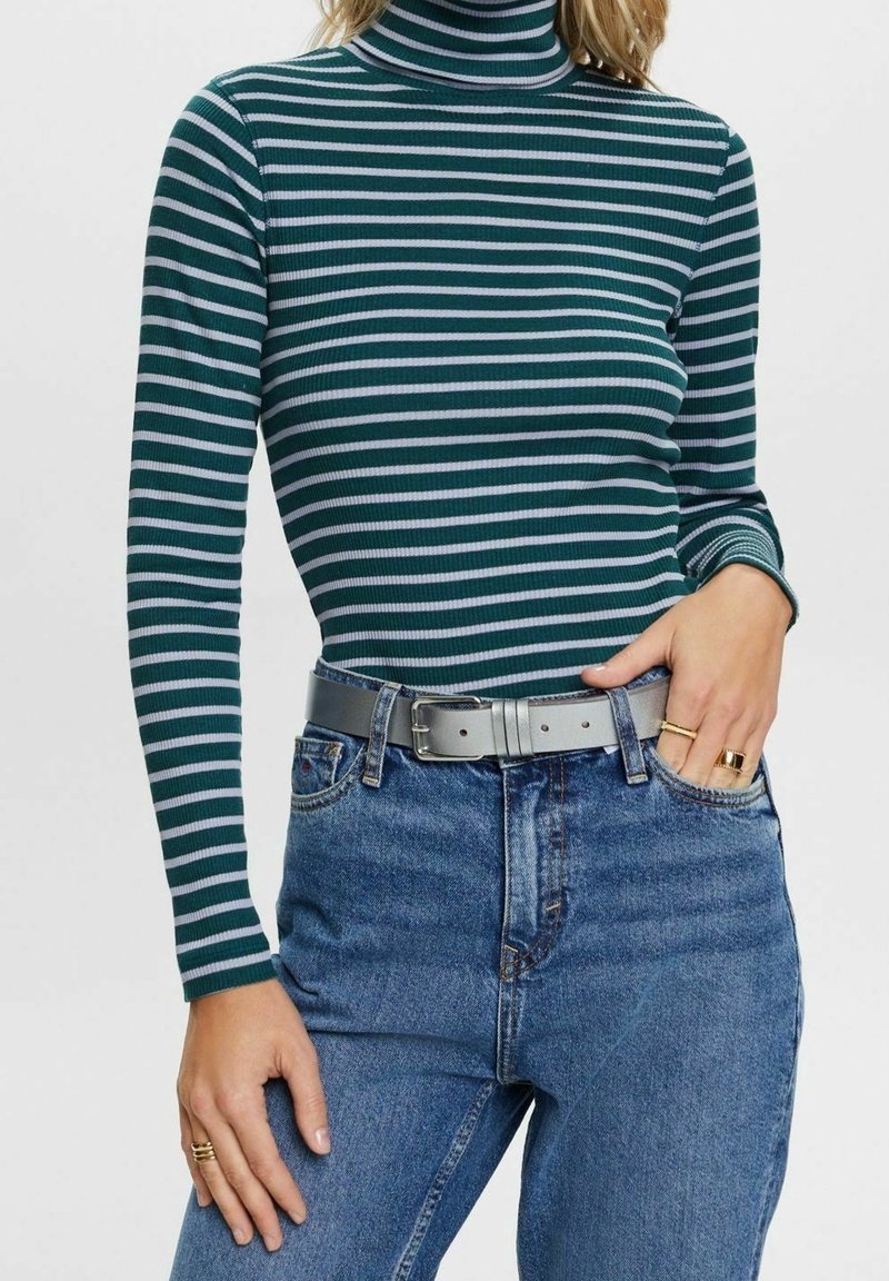 Femme portant un col roulé rayé vert et blanc, un jean taille haute bleu avec une ceinture grise, et des bagues en or à la main gauche.