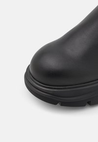Gros plan sur le bout d'une chaussure en cuir noir avec une semelle épaisse en caoutchouc et une bande de roulement texturée pour une bonne adhérence sur un fond clair uni.