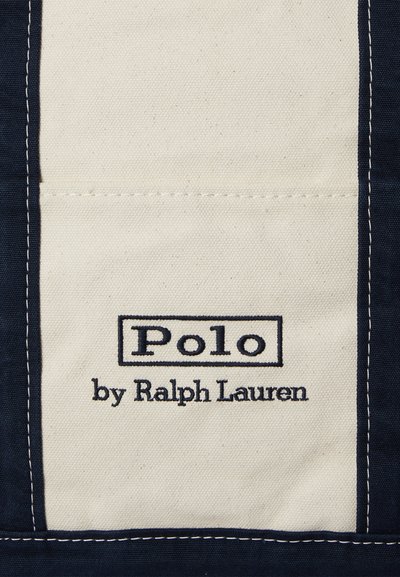 Vászon címke tengerészkék és krémszínű csíkokkal. A szöveg „Polo” vastag betűkkel, alatta kisebb betűmérettel „by Ralph Lauren” felirattal.