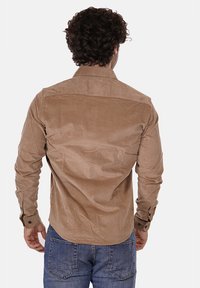 Camicia in corduroy marrone con colletto, maniche lunghe e dettagli con bottoni sui polsini. Tessuto liscio e vestibilità aderente. Indossata con jeans blu.