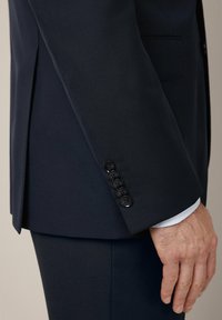 Granatowa marynarka o gładkiej teksturze, z jednorzędowym zapięciem i sześcioma czarnymi guzikami na mankiecie. Biała koszula widoczna poniżej.