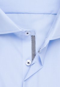 Camicia azzurro chiaro con collo classico, bottoni bianchi e motivo pied de poule bianco e nero lungo la patta interna.