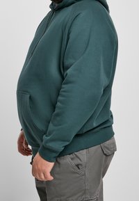 Homme portant un sweat à capuche bleu sarcelle et un pantalon cargo gris, vu de profil sur un fond clair uni.