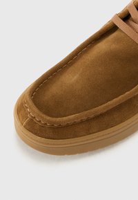 Chaussure en cuir suédé marron avec un bout arrondi, une semelle en caoutchouc et des coutures contrastées. Elle présente un laçage en haut pour un ajustement sécurisé.