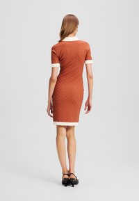 Terrakotové bodycon šaty s texturovaným mřížkovým vzorem, bílým límečkem a manžetami, končící nad koleny. Černé podpatky dokončují vzhled.