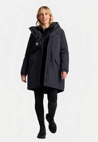 Marineblauwe lange parka met ritssluiting en knopen, grote zakken, verstelbare capuchon en elastische manchetten, gedragen over zwarte leggings en sneakers.