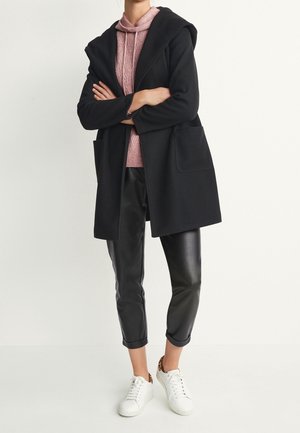 Personne portant un manteau noir par-dessus un sweat à capuche rose, un pantalon en cuir noir coupé court et des baskets blanches avec des accents de talon imprimé léopard.