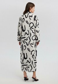 Abito lungo bianco con motivi astratti neri. Presenta maniche lunghe e un colletto, con una linea dritta e un tessuto fluido. Sandali neri con tacco.