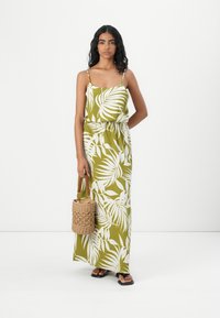 Robe maxi vert olive avec motif de feuilles blanches, bretelles spaghetti, taille élastique, associée à un sac en osier et des sandales noires.