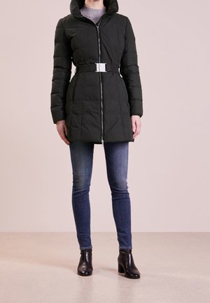 Kobieta w czarnej pikowanej kurtce z paskiem, niebieskich dżinsach i czarnych botkach, stojąca na tle jednolitego beżowego tła.