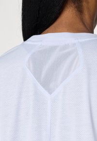 Bílý sportovní tričko s mřížkovaným vložením na zadní straně, se vzorem ve tvaru V. Hladká textura s malými perforacemi po celém povrchu.
