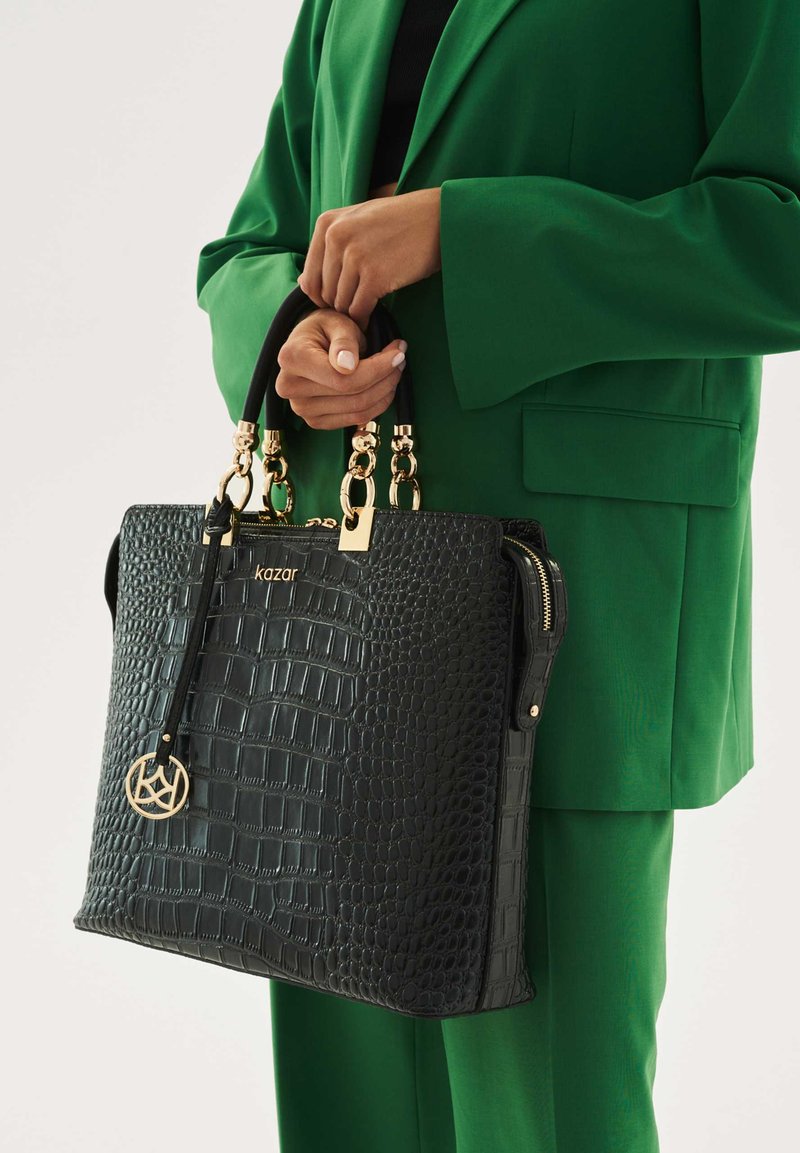 Musta krokotiilikuviollinen käsilaukku, jossa on kultaiset metalliosat, kaksinkertaiset yläkahvat ja irrotettava koru. Pinta on teksturoitu ja laukku on jämäkän rakenteinen.