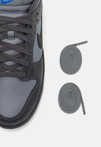 Sive Nike športni copat z sintetičnim zgornjim delom, mrežastimi paneli in črnimi poudarki. Dve okrogli sivi vezalki sta prikazani ob čevlju.