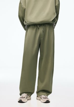 Olive green loose-fit sweatpants with drawstring waist paired with white and gray chunky sneakers on a plain background.