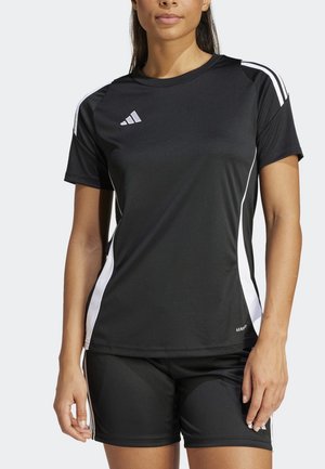 Femme portant une tenue de sport Adidas noire avec des bandes blanches, comprenant un t-shirt à manches courtes et un short, debout les bras détendus.