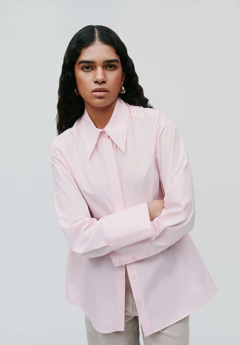 Camicia button-up rosa chiaro con colletto a punta, maniche lunghe e vestibilità ampia, con orlo dritto e una sottile pettorina con bottoni frontali.
