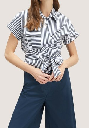 Kvinde iført kortærmet bluse med lodrette marineblå og hvide striber, med sløjfe foran og lomme, kombineret med marineblå bukser med vide ben.