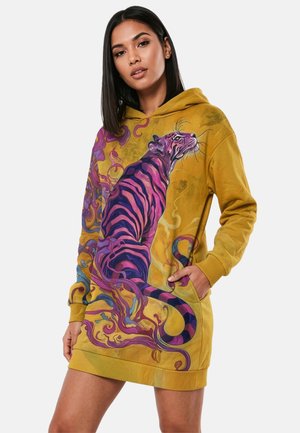 Junge Frau trägt ein senfgelbes Kapuzenkleid mit großem lila-rosa gestreiftem Tigerbild und wirbelnden abstrakten Mustern.