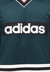 Zöld hálós sportmez V-nyakkal, nagy fekete téglalappal, amelyben fehér, félkövér "adidas" felirat található, valamint fehér és fekete vízszintes csíkok a szegélyén.