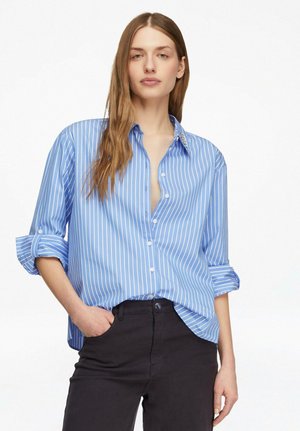 Młoda kobieta z długimi włosami, ubrana w niebiesko-białą koszulę w paski z podwiniętymi rękawami oraz czarne spodnie, pozująca z jedną ręką w kieszeni.