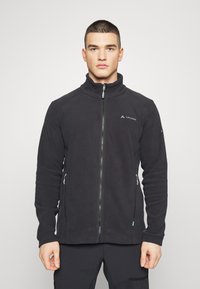 Schwarze Fleecejacke mit Stehkragen, frontaler Reißverschluss, zwei seitlichen Reißverschlusstaschen und Logo auf der linken Brust. Glatte Textur.