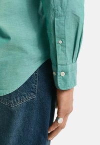 Helles grünes Hemd mit einer strukturierten Oberfläche, mit einem spitzen Kragen und Manschetten. Kombiniert mit dunkelblauen Jeans und einem silbernen Ring.