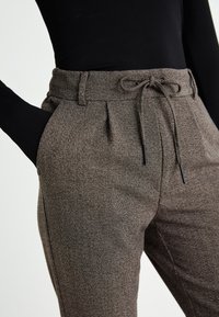 Pantaloni marroni in tessuto testurizzato con vita regolabile con coulisse, pieghe frontali e tasche. Realizzati in un tessuto intrecciato con un motivo sottile.