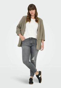 Blazer oversized en verde oliva, camiseta blanca, jeans ajustados desgastados en gris y zapatillas negras con detalles en blanco. Conjunto informal con texturas suaves.