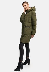 Olivgrön vadderad parkas med huva, med dragkedja fram, flera fickor och ett quiltat mönster. Bärs med svarta skinny jeans och ankelstövlar.