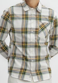 Mann trägt ein weißes Hemd mit Knöpfen, das ein kariertes Muster in Grün, Gelb, Braun und Grau aufweist, mit einer Brusttasche und Kragen.