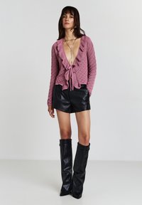 Donna che indossa un top in maglia rosa con scollatura a V profonda e volant, pantaloni corti di pelle nera, stivali alti neri di pelle e lunghe collane di argento a strati.