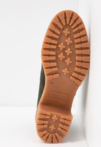 Suela de goma marrón con un patrón de superficie texturizado, que presenta agarres elevados y una sección plana. La parte superior es de un material verde oscuro con detalles de costura.