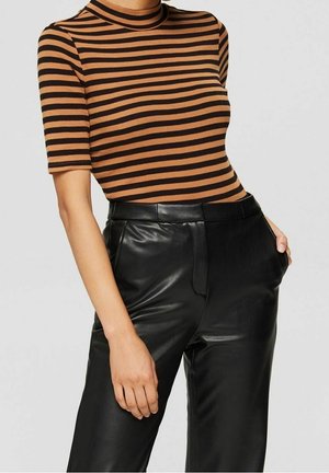 Mujer con una camiseta de manga corta con rayas horizontales marrones y negras, metida en unos pantalones de cuero negros de talle alto, con las manos en los bolsillos.