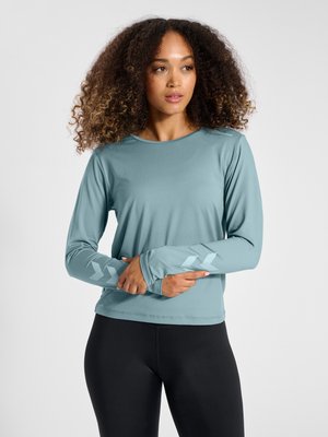 Woman with curly hair wearing a light blue long-sleeve shirt with arrow designs on sleeves and black leggings, facing slightly right.