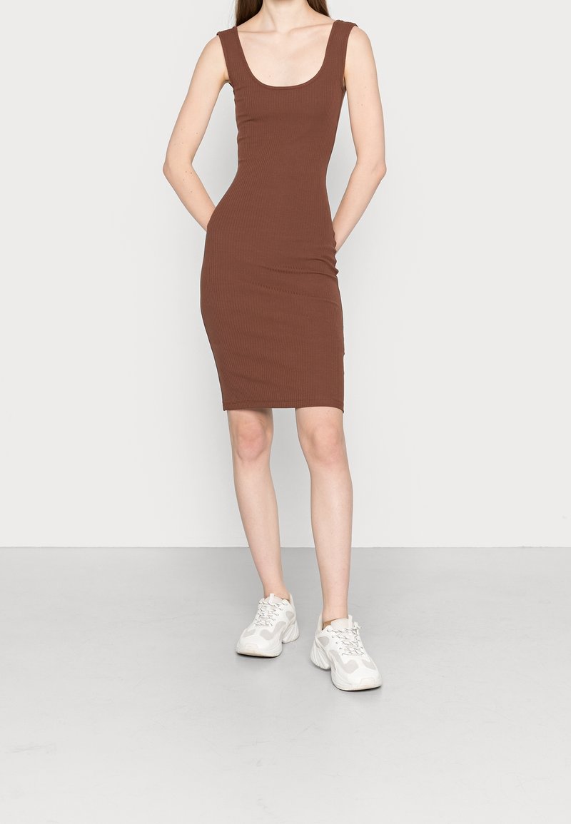 Abito canotta marrone a costine con scollatura a giro, silhouette aderente e lunghezza al ginocchio. Il modello indossa sneakers bianche. Sfondo minimalista.